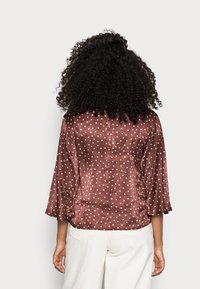 Brun satinblus med vita prickar, med 3/4 ärmar och en lös passform. Slät textur med en subtil lyster.