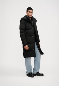 Casaco puffer preto com capuz, apresentando um fecho zip e de pressão, grandes bolsos e comprimento até a metade da coxa. Combinado com jeans azuis e sapatos pretos.
