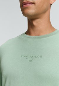 Felpa in cotone verde con colletto a girocollo, con logo 'TOM TAILOR' tono su tono nella zona del petto. Texture liscia e design casual.
