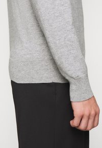 Maglione grigio a maglia con scollatura rotonda, polsini e orlo a coste. Indossato con pantaloni neri, evidenziando la texture e la vestibilità.