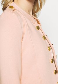 Cardigan rose avec un col rond, des boutons dorés ornant l'épaule et le devant, et un tissu doux et texturé.