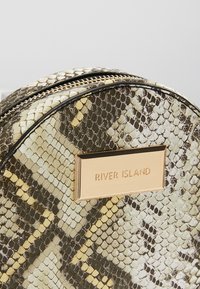 Sac à main rond avec motif en peau de serpent aux tons beige, marron et jaune, avec une plaque métallique dorée portant le texte "River Island".