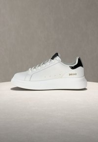 Λευκό sneaker χαμηλού τύπου με μαύρη έμφαση στη φτέρνα, κορδόνια εμπρός, παχύ πάτο και λογότυπο "Just Cavalli" στην πλευρά, εκτεθειμένο σε ουδέτερη επιφάνεια.