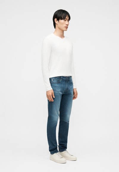 Νεαρός άνδρας σε όρθια στάση, φορώντας λευκό πουκάμισο με μακριά μανίκια, μπλε τζιν και λευκά αθλητικά παπούτσια, μπροστά σε απλό φωτεινό φόντο.