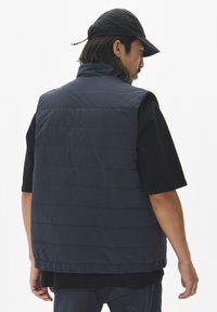 Navy blue puffer vest with vertical stitching, high collar, and elastic hem, worn over a black short-sleeve shirt.