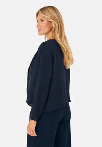 Marineblauer Cardigan aus strukturiertem Stoff, offene Front, 3/4-Ärmel und lockere Passform, getragen über dunklen Hosen.