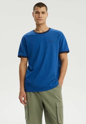 Młody mężczyzna stoi z rękami w kieszeniach, ubrany w niebieską koszulkę z ciemnymi wykończeniami i oliwkowozielone spodnie cargo na jednolitym jasnym tle.