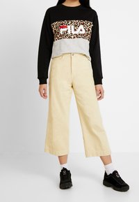 Osoba ubrana w czarno-szarą bluzę FILA z panterkowym nadrukiem, beżowe spodnie o szerokich nogawkach, białe skarpetki i czarne trampki stoi na białej podłodze.