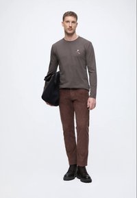 Rudas ilgomis rankovėmis marškinis su siuvinėjimu, rudi zomšiniai kelnių, juodi batai ir juodas petis krepšys. Minimalistinis dizainas ir pritaikymas.