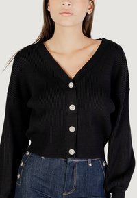 Cardigan nero a costine con scollatura a V e cinque bottoni in perla. Maniche lunghe e design corto. Abbinato a jeans in denim scuro.