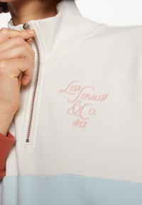 Vit zip-up sweatshirt med pastellblå och korallfärgade accenter. Har en broderad rosa logotyp "Levi Strauss & Co." med en slät textur och ribbad krage.