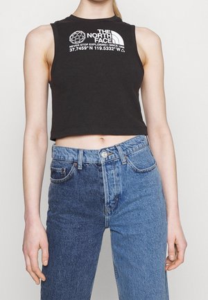 Nainen, jolla on musta, lyhyt hioton toppi, jossa on "The North Face" -logo, ja siniset kaksisävyiset korkeavyötäröiset farkut, seisoo yksiväristä taustaa vasten.