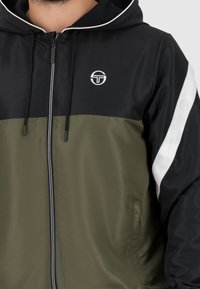 Schwarz und olivgrün Kapuzenjacke mit Reißverschluss, kontrastierenden weißen Akzenten und Markenlogo auf der Brust und dem Ärmel. Texturierte Stoffdetails sind enthalten.