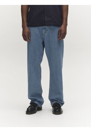 Férfi laza kék farmerben, fekete bőr masnis loaferekben és fekete gombos ingben, áll egy egyszerű világos háttér előtt.