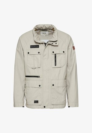 Beige udendørs jakke lavet af tekstureret stof, med fire lommer, en lynlås og en stående krave. Sorte accenter fremhæver designet.