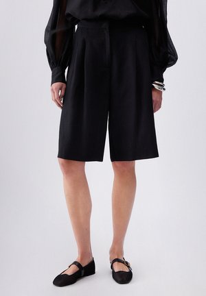 Persoon draagt zwarte knielange wijde korte broek, zwarte mesh Mary Jane platte schoenen, doorschijnende zwarte top met lange mouwen en zilveren armbanden.