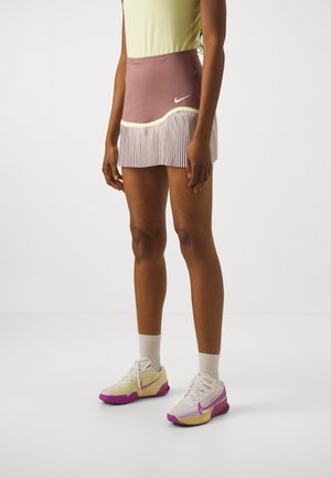 Kvinde iført en lilla Nike plisseret tennisnederdel med gul kant, lysegul top, hvide sokker og hvide og lilla Nike sneakers.