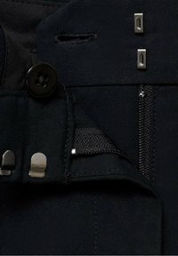 Détail d'un vêtement en tissu noir avec un bouton, des fermoirs métalliques et une ouverture zippée, finition texturée et poche intérieure.