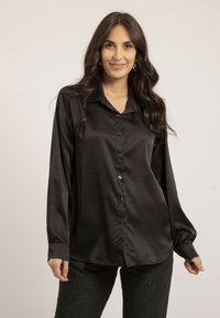 Chemise en satin noir avec col, manches longues et boutons sur le devant. Le tissu a une texture lisse, associé à un pantalon sombre et ajusté.