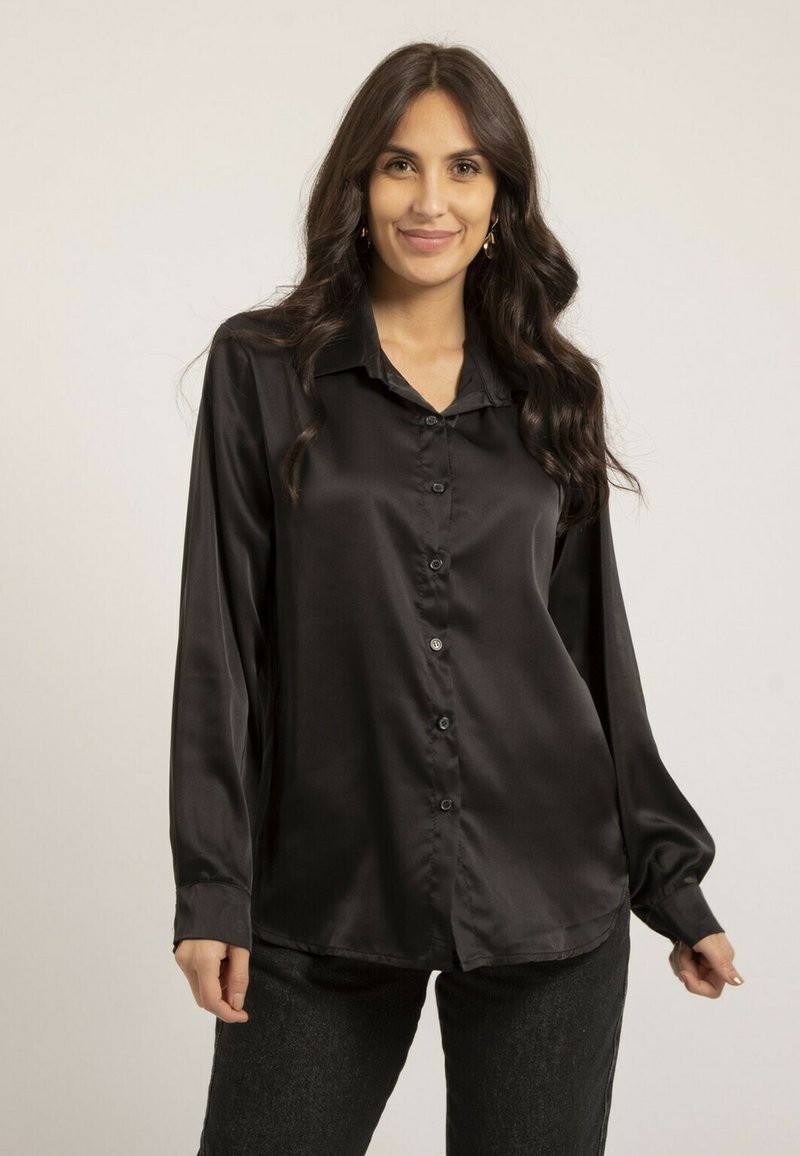 Chemise en satin noir avec col, manches longues et boutons sur le devant. Le tissu a une texture lisse, associé à un pantalon sombre et ajusté.