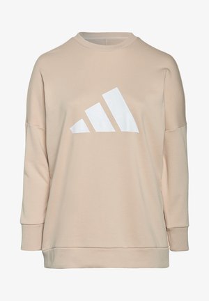 Personne portant un sweat-shirt beige à manches longues avec un grand logo blanc Adidas, associé à un pantalon foncé, recadrée à la tête et aux genoux.