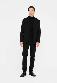 Schwarzer Anzug mit einem Zweiknopfjackett, schmal geschnittenen Hosen und einem schwarzen Rollkragenpullover darunter. Die Schuhe sind aus glänzendem schwarzem Leder mit einem eleganten Design.