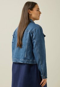 Jeansjacka i ljusblått med klassisk skärning, med knapppdetaljer och en något kortare design. Stylistiskt över en mörkblå klänning.