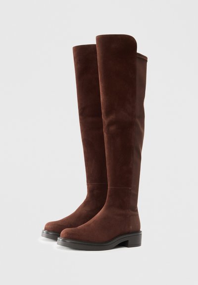 Alte stivali in suede marrone scuro con tacco basso e punta arrotondata, in piedi affiancati su uno sfondo bianco.