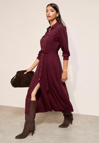 Dlhé vínové šaty košieľkového strihu s predným zapínaním na gombíky, priviazaným pásom, dvomi prsnými vreckami a vysokým rozparkom. Nosia sa s hnedými semišovými čižmami po kolená.