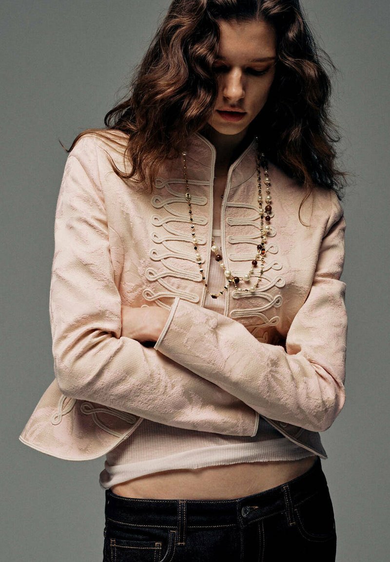 Jeune femme aux longs cheveux ondulés porte une veste décorative rose pâle, un collier de perles et un jean foncé, debout les bras croisés et regardant vers le bas.