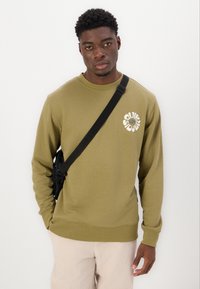 Olivovo zelená mikina s okrúhlym výstrihom a dlhými rukávmi, s bielym grafickým logom na hrudi. Nosí sa s béžovými nohavicami a čiernou crossbody taškou.