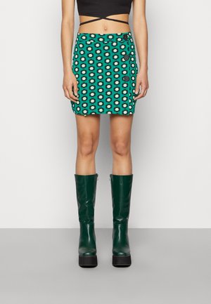 Saia mini de padrão verde com desenhos florais, cintura alta e detalhes de botões, combinada com botas verdes até ao joelho e uma blusa preta curta.