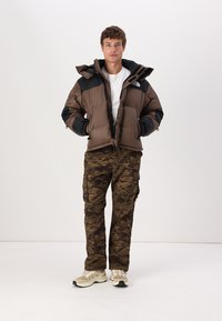 Brun puffjacka med svarta accenter, dragkedjefickor och huva. Kombination med kamouflage cargo-byxor och beige sneakers.