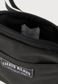 Μαύρη ύφαντη τσάντα με λευκό φερμουάρ. Το εσωτερικό διαθέτει γκρι ετικέτα με το λογότυπο «Calvin Klein». Η επιφάνεια έχει λεία υφή και ορατή ραφή.