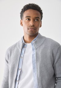 Grå, rillede cardigan lagt over en lys blå skjorte med knapper, båret over en hvid t-shirt. Har høj krave og træknapper.