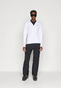 Biała bluza z kapturem zapinana na zamek, na czarnej koszulce, w połączeniu z czarnymi spodniami narciarskimi i czarnymi butami narciarskimi. Akcesoria obejmują czarne okulary przeciwsłoneczne.