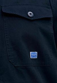 Mørkeblå skjortelomme med knappet klaff og liten blå rektangulær Jack & Jones Premium-label sydd nær nederste kant.