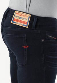 Κοντινό πλάνο ατόμου που φοράει σκούρα μπλε τζιν Diesel με καφέ ετικέτα και κόκκινη ραφή λογότυπου στην πίσω τσέπη.