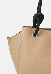 Beżowa skórzana torba tote o teksturowanym wykończeniu, z czarnym uchwytem, srebrnymi detalami w postaci oczek i otwartą konstrukcją.