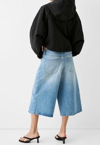 Klippt svart hooded jacka i kombination med ljusblå vida denim culotter, med fransade fållar och bakfickor, kompletterat med svarta sandaler.