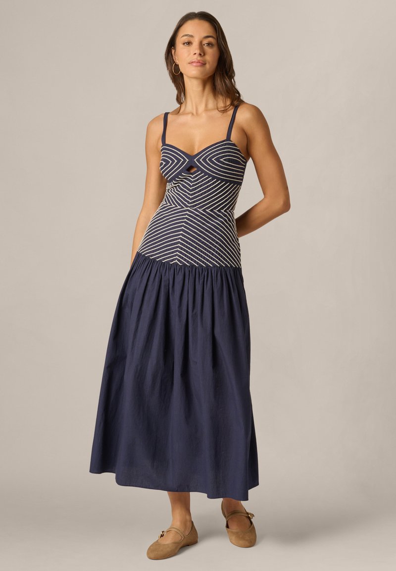 Frau steht in einem marineblauen Maxikleid mit weißen diagonalen Streifen am eng anliegenden Oberteil und beigefarbenen flachen Schuhen vor einem einfarbigen Hintergrund.
