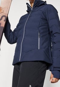 Personne portant une veste d'hiver zippée bleu marine et un pantalon noir, tenant un snowboard avec un design rose et noir sur le côté gauche.