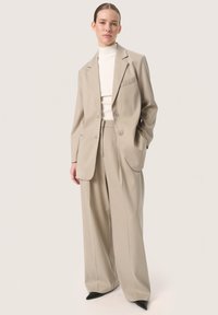 Beige skræddersyet blazer med to knapper, enkelt brystlomme og vide benbukser, parret med en tætsiddende hvid rullekrave og sorte sko.