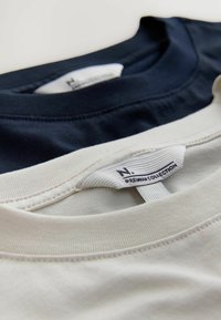 Trois t-shirts en bleu marine, blanc et beige clair ; en coton, avec des encolures rondes et des étiquettes de marque visibles cousues à l'intérieur.