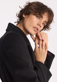 Młoda osoba z krótkimi brązowymi włosami i niebieskimi oczami, ubrana w czarny płaszcz, patrzy w kamerę z dłońmi delikatnie złożonymi blisko twarzy.