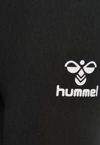 Biała stylizowana etiuda pszczeli z napisem "hummel" na czarnej tkaninie, prawdopodobnie część odzieży sportowej.