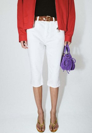 Persona que lleva pantalones blancos recortados con un cinturón trenzado marrón, chaqueta roja, sandalias doradas y sostiene un bolso pequeño morado.