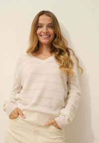 Jasny beżowy sweter w serek z poziomymi paskami, o miękkiej teksturze, luźnym kroju, oversizedowych rękawach i prążkowanych mankietach. Kieszenie widoczne w jasnych spodniach.