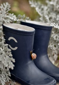 Kék gumicsizma sima textúrával és világos béléssel. Egy csíkos csiga pihen az egyik csizmán. Körülötte zöld lombok.