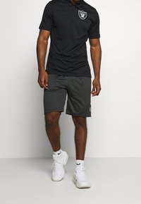 Mies kävelee päällään musta Raiders-polo-paita, tummanharmaat shortsit, valkoiset Nike-sukat ja valkoiset lenkkarit vaalealla taustalla.